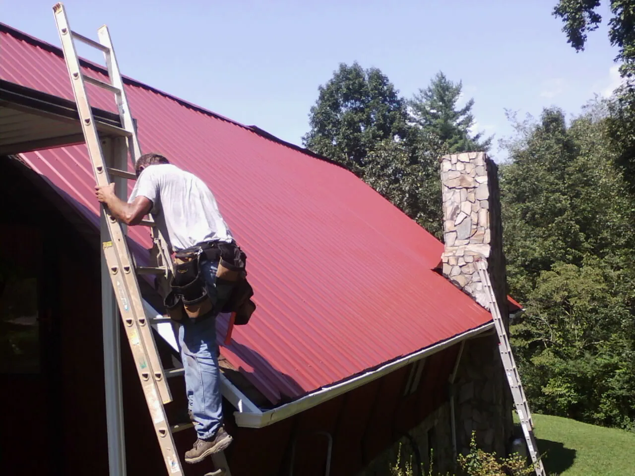 Roofer on a ladder beside a red metal roof and stone chimney — Soffit & Fascia Repair by Roofing Brothers in Onaga